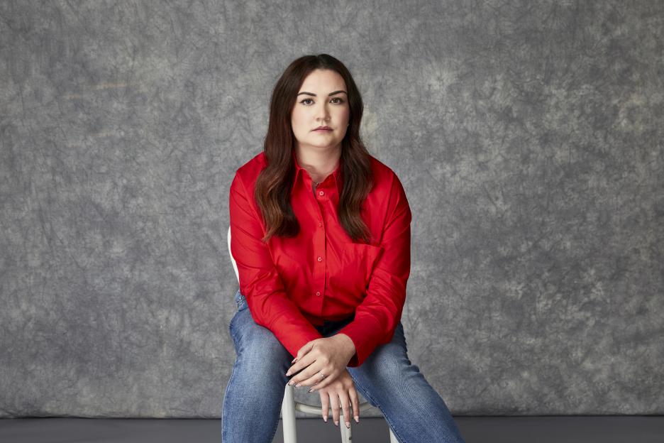 Lauren Eden Pen wearing a red shirt and jeans, sitting on a white chair against a gray background.
