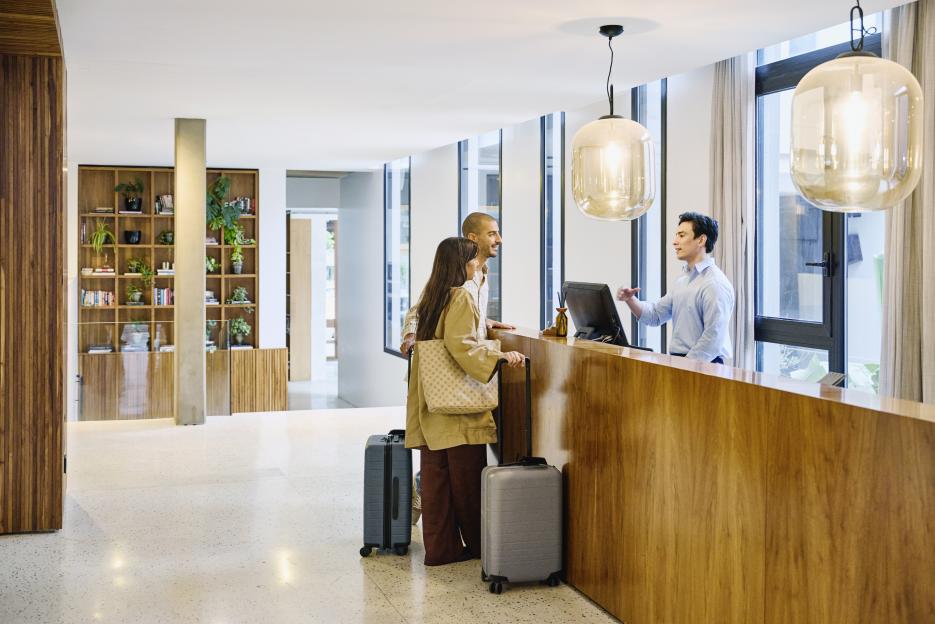Male hotel receptionist assisting a couple with suitcases at the front desk.