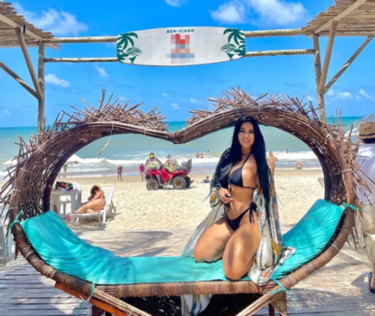 A woman in a bikini poses in a heart-shaped chair on a beach with a blue sky.