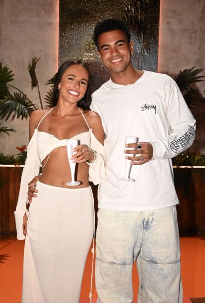 Two people, a woman and a man, smiling and holding champagne flutes at the 'Love Island: Aftersun' TV Show.