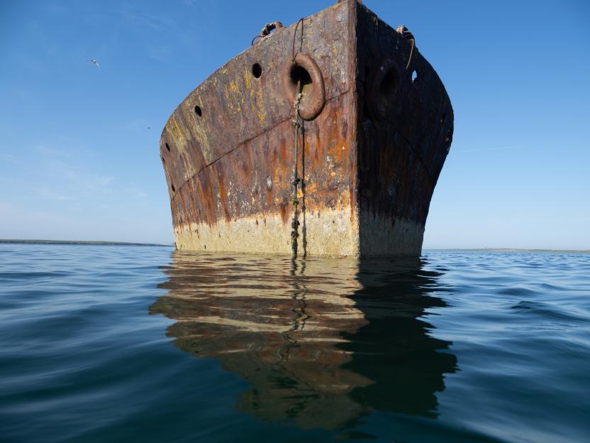 A rusted, derelict ship's hull partially submerged in calm blue water with a bright blue sky overhead.