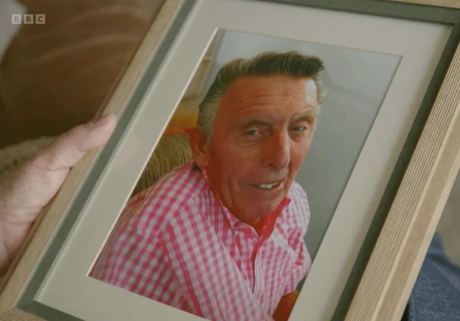 A person holding a framed photo of a smiling man in a pink and white checkered shirt.