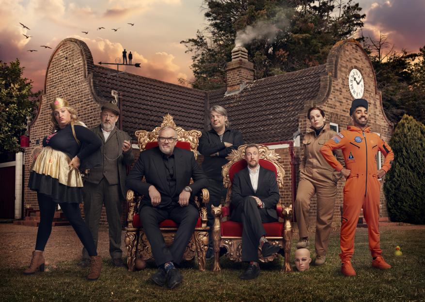 A diverse group of seven comedians posing in front of a house at sunset, including Greg Davies, Daisy May Cooper, Jonny Vegas, Richard Herring, Alex Horne, Katherine Parkinson, and Mawaan Rizwan.