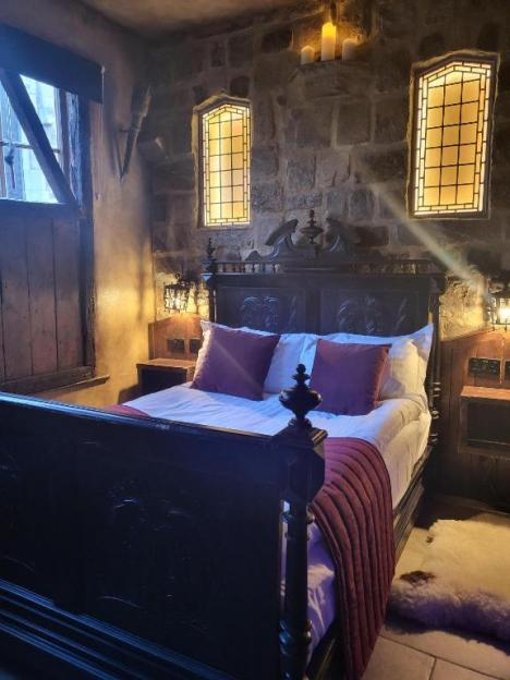 A dimly lit hotel room in a historic building featuring a dark wooden bed with white and red bedding, stone walls, and two stained-glass windows.