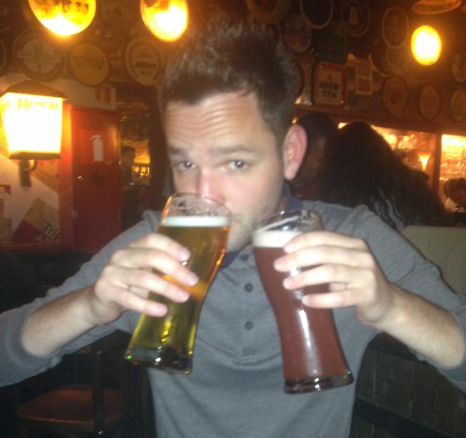 A man in a grey shirt and blue jeans holding a glass of pale beer and a glass of dark beer.