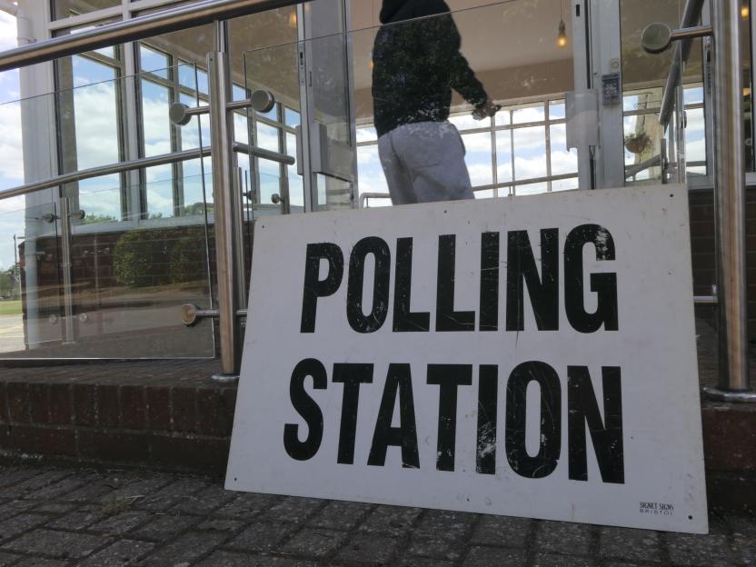 A polling station sign on the ground with a person in a tracksuit entering a building in the background.