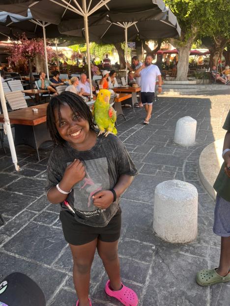 A young girl smiling at the camera with a parrot perched on her shoulder at an outdoor restaurant.