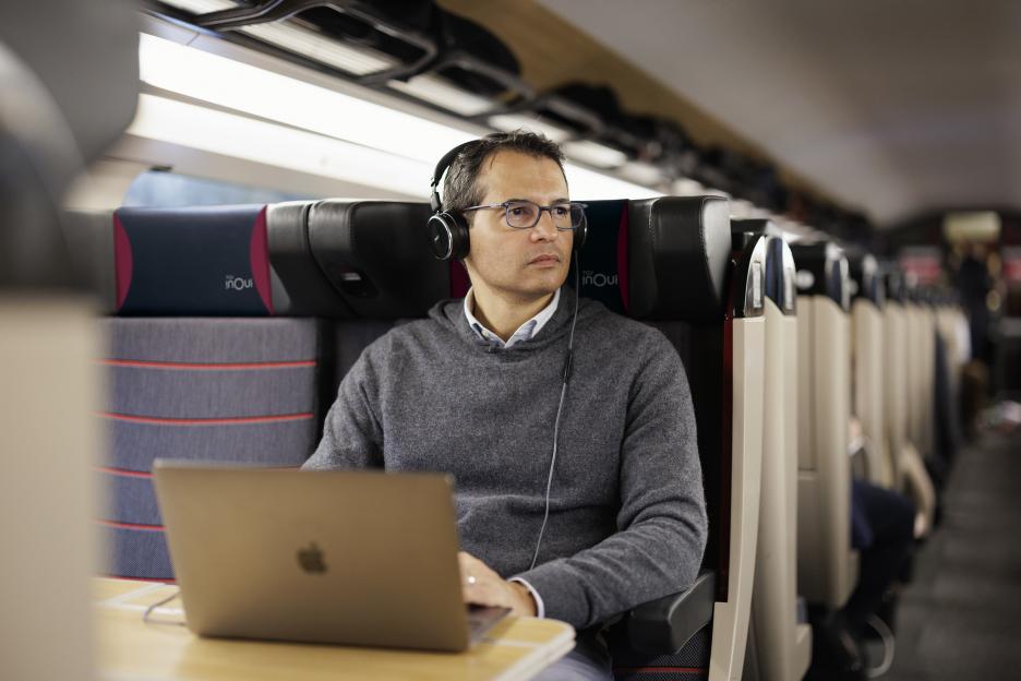 A man wearing headphones and glasses works on a laptop in an OPTIMUM TGV InOui train car.