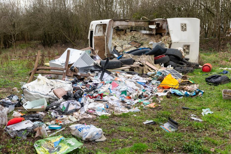 A fly-tipping site with a burnt-out caravan, tires, and rubbish bags.