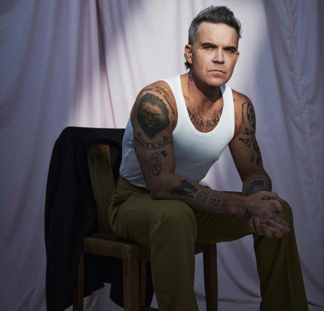 Robbie Williams seated, wearing a white tank top and olive green pants, looking directly at the camera.