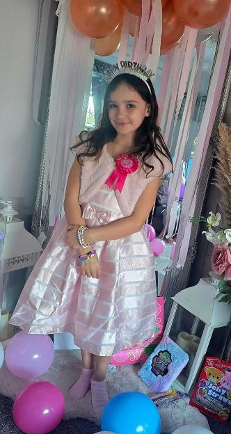 Aria Thorpe, a young girl, smiles wearing a pink party dress, a birthday crown, and colorful bracelets.