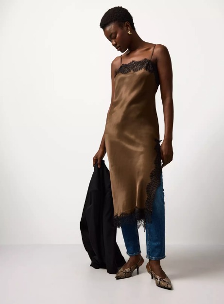 A woman models a brown satin midi dress with black lace trim over blue jeans, holding a black jacket, wearing snakeskin heels.