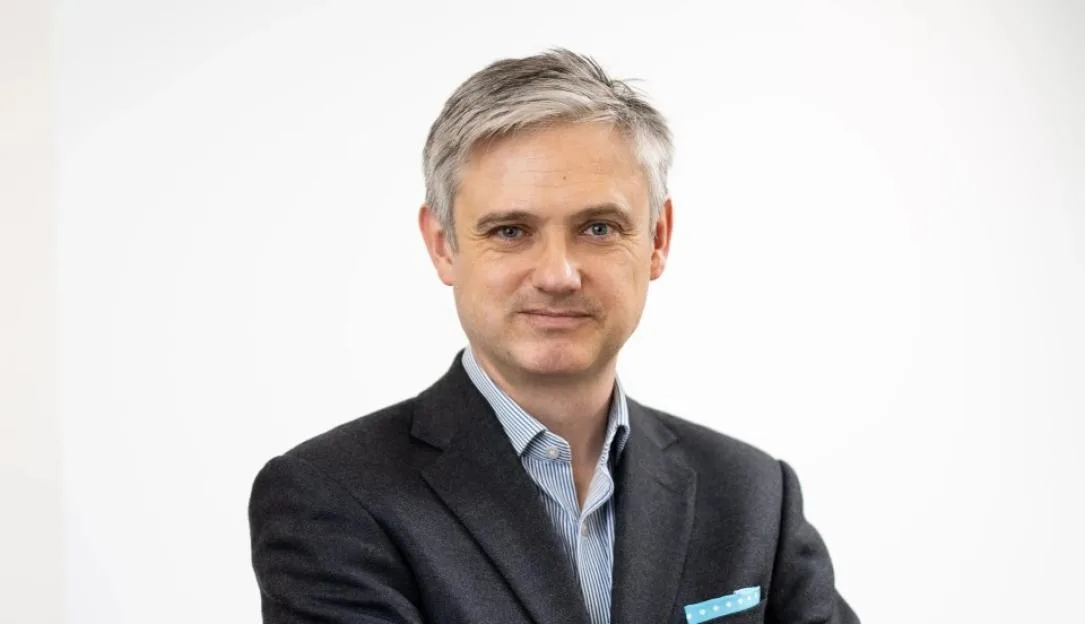 Lord Walney stands with his arms crossed, wearing a grey suit jacket, blue and white striped shirt, and a light blue polka-dotted pocket square.