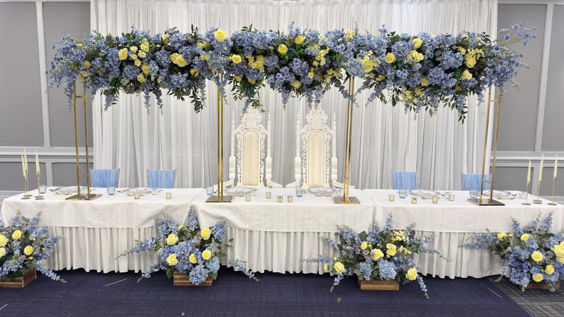 A wedding reception room with a top table adorned with blue and yellow flowers, featuring two ornate thrones.