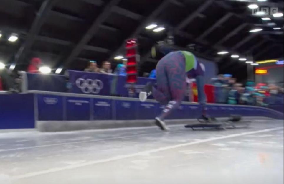 A skeleton racer in a purple and green suit crouches, preparing to push their sled across the ice.