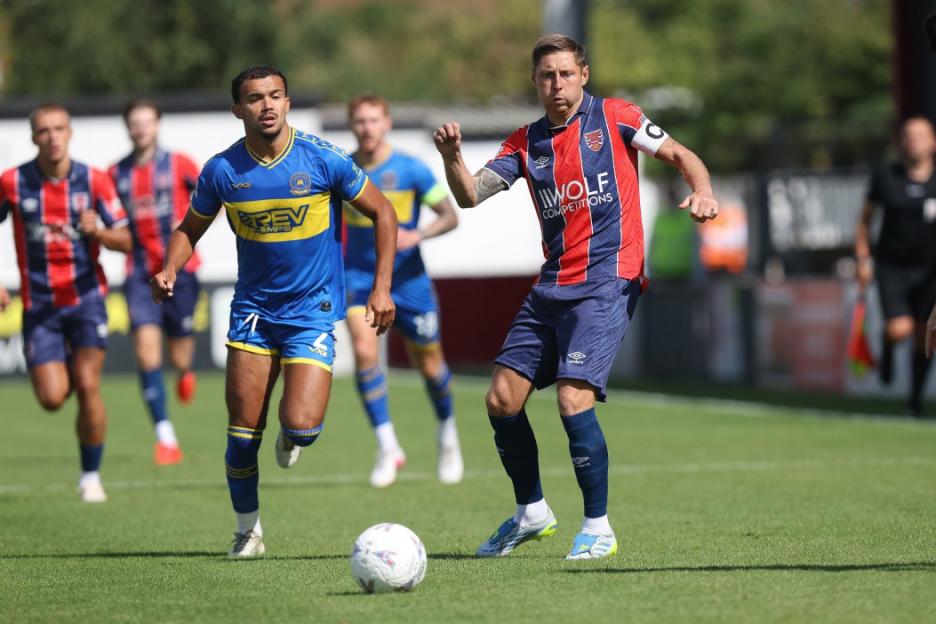 Two footballers, one in blue and yellow and the other in red and blue, are on the field during the Dagenham & Redbridge vs Torquay United match.