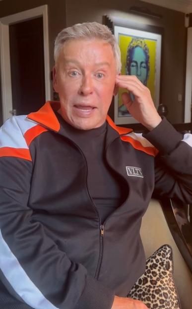 Man with fair skin and light gray hair wearing a black, orange, and white tracksuit.