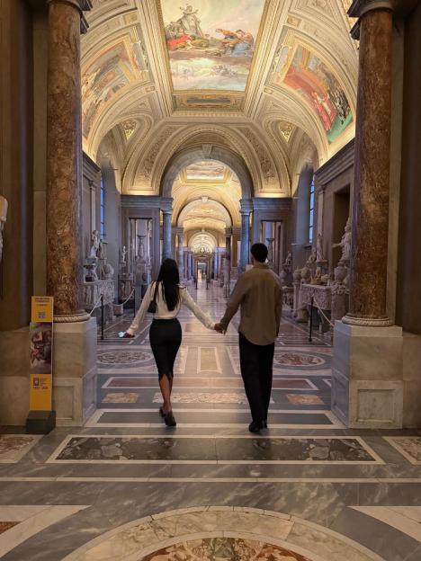Maya Jama holding hands with a man, walking away in a grand hall with painted ceilings and statues.