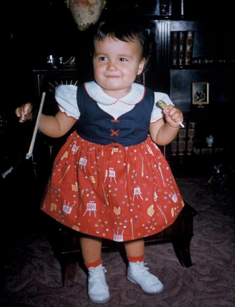 A young Diana wearing a red dress with white artist's easels printed on it, holding a paintbrush and a lollipop.