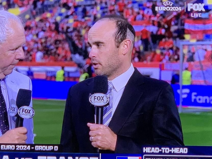A man with a receding hairline speaking into a FOX Sports microphone next to another man, both broadcasting from a stadium.