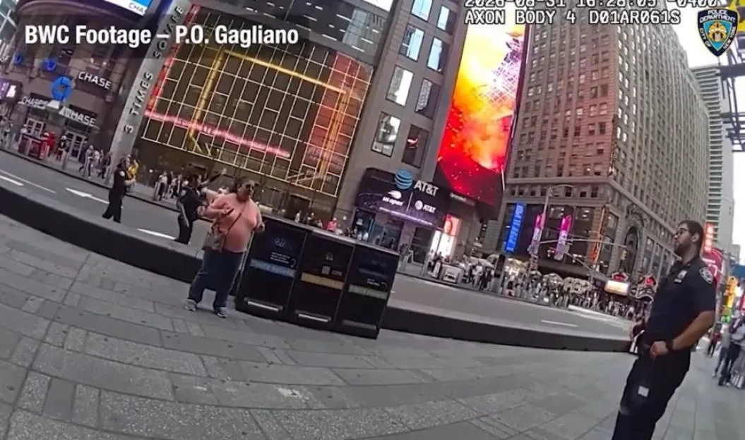 Bodycam footage of a New York City police officer standing on a sidewalk in Times Square, with a woman walking past and buildings and other people in the background.