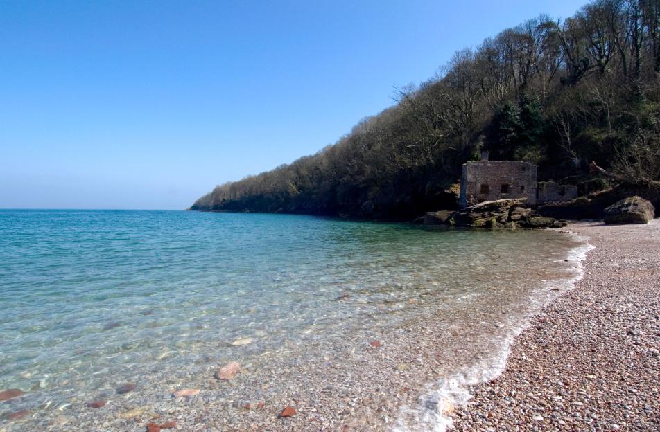 Elberry Cove, a rocky beach with clear blue water and an old stone building at the base of a wooded hill.