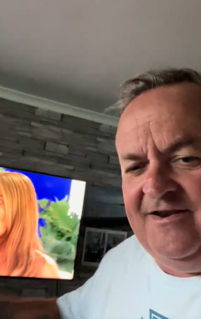 A man filming himself with a TV showing a woman in the background.