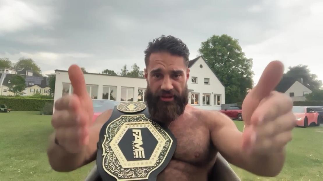 A man with a beard and a championship belt with "FAME FIGHTING" on it gesturing with both hands.