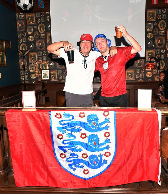 Conah Mckenna and Daniel Cadden smiling at the camera, holding drinks, with an England flag draped in front of them.