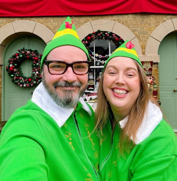 A couple dressed in green elf outfits and hats smiling in front of a heavily decorated Christmas home.
