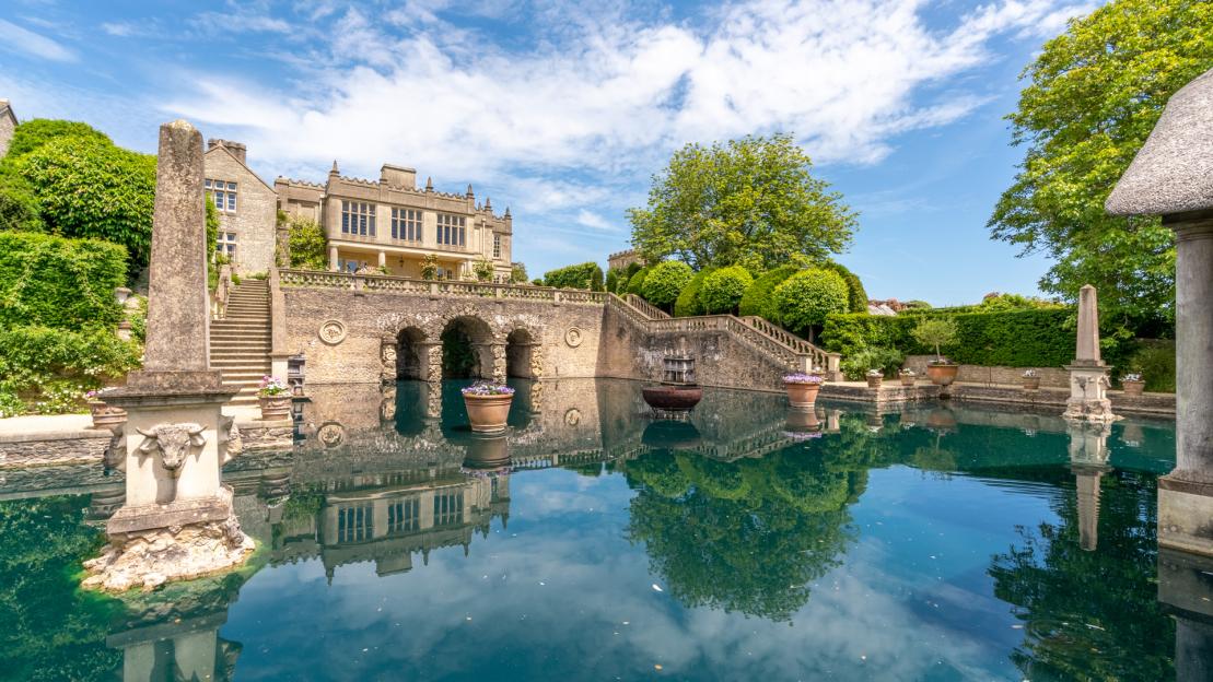 The Euridge Manor in Wiltshire, with its large manor house and stone arches reflected in the blue water of a pond.