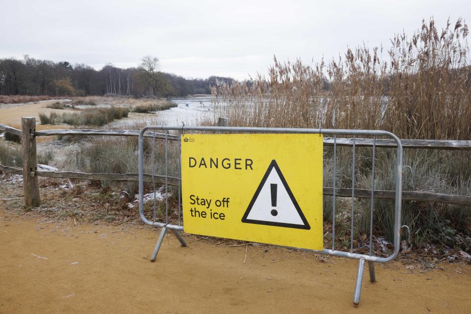 A yellow "DANGER Stay off the ice" sign on a metal barrier in front of Pen Ponds in Richmond Park.