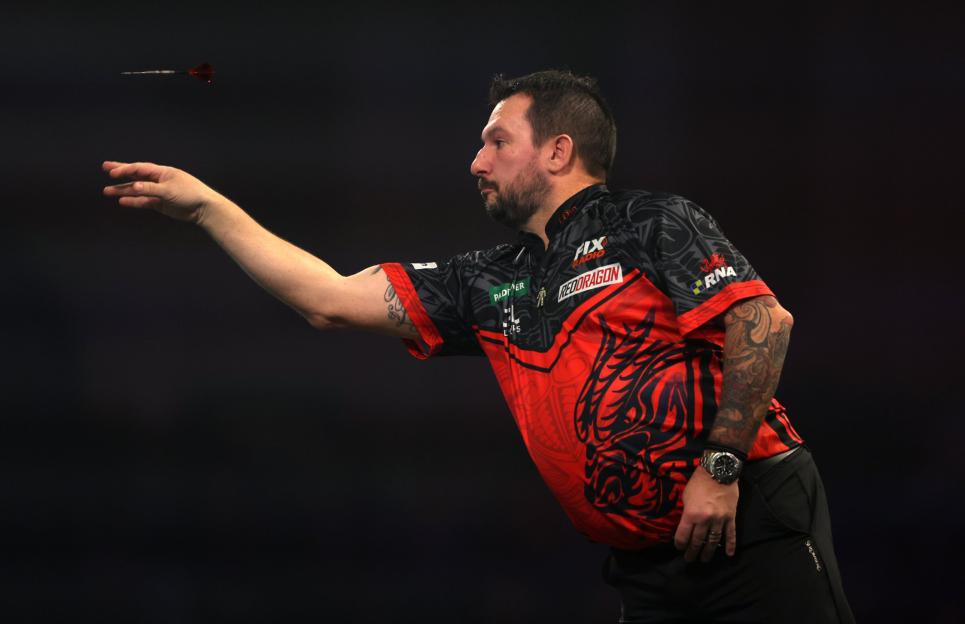 A darts player in a red and black shirt throws a dart.