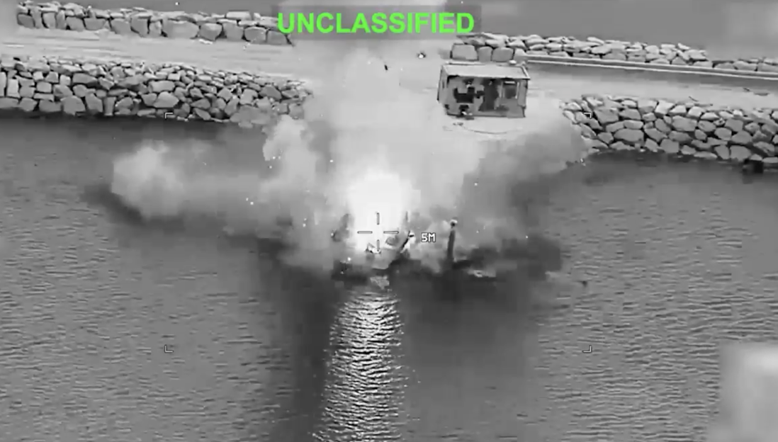 Drone footage shows an explosion on a small boat in the water next to a rocky shoreline with a small building.