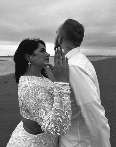 Black and white photo of a woman in an elaborate pearl-embroidered top and skirt, looking up at a man in a white shirt, showing off her engagement ring.