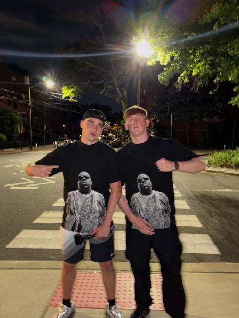 Two young men wearing black t-shirts with a large image of The Notorious B.I.G. on them, standing on a crosswalk at night.