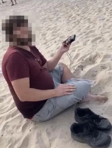A man sits cross-legged on the sand at a beach, looking at a device in his hand, with his face blurred for privacy.