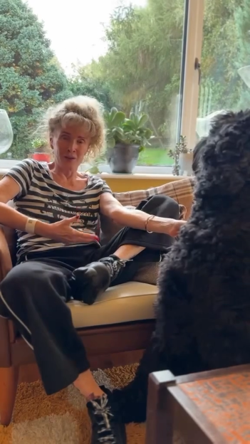 Beverley Callard sitting on a couch with a large black dog, wearing a hospital wristband.