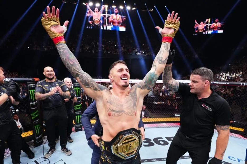 UFC fighter Tom Aspinall raises his arms in victory, wearing gold gloves and a championship belt, with a referee beside him and a crowd in the background.