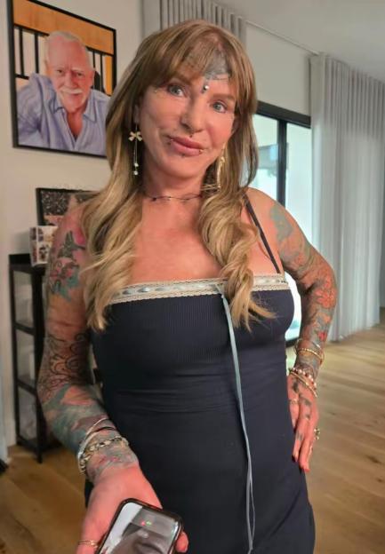 A woman with tattoos on her arms and forehead, and facial piercings, holds a phone.