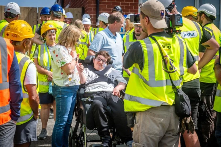 A young person in a wheelchair is surrounded by family and building crew members in hi-vis vests.
