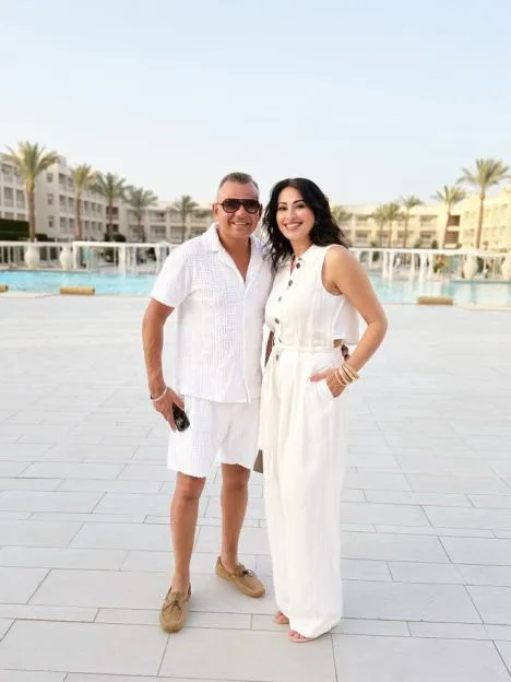 Jinda Mann and her husband, Bindu, pose together outdoors in matching white outfits near a pool and resort buildings.