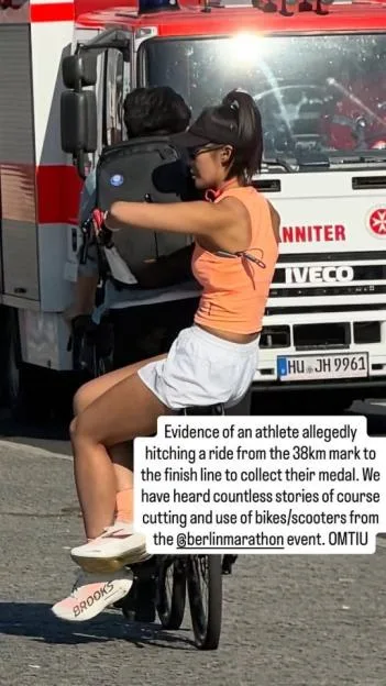 A runner in an orange top and white shorts rides on the back of a bicycle past a white and red vehicle.