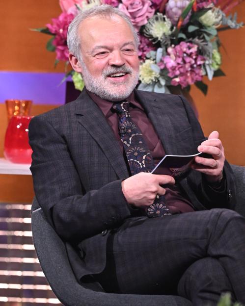 Graham Norton sitting and smiling, wearing a dark suit and patterned tie.
