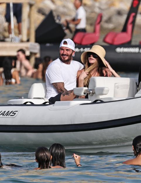 David and Victoria Beckham in a boat on the water, with swimmers nearby.