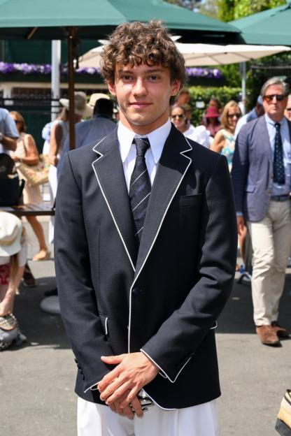 A young man in a dark blazer with white trim and a striped tie looks at the camera.