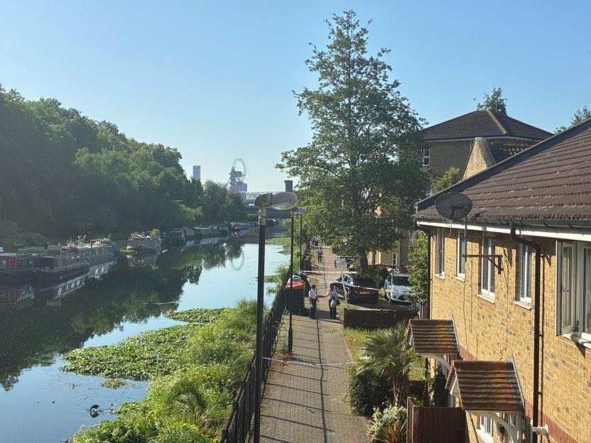 Police tape blocking off a canal path with emergency personnel and vehicles, with boats docked and buildings nearby, and the London Eye visible in the distance.