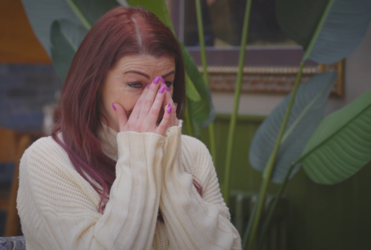 A woman with red hair and purple nail polish covers her face with her hands, crying tears of joy.