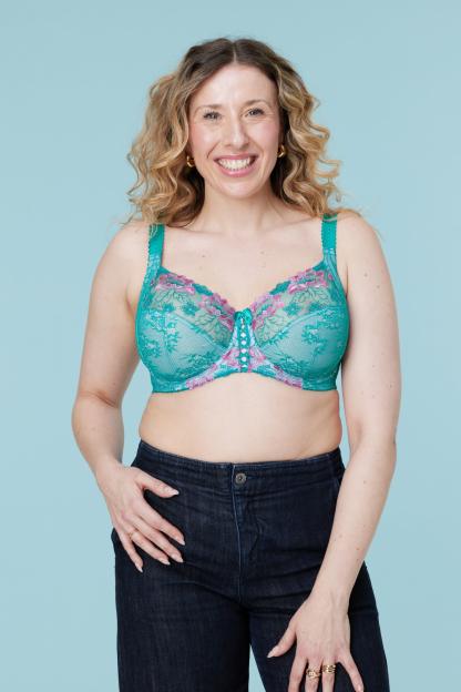 A smiling woman with curly blonde hair wears a teal lace bra with pink floral embroidery and dark jeans.