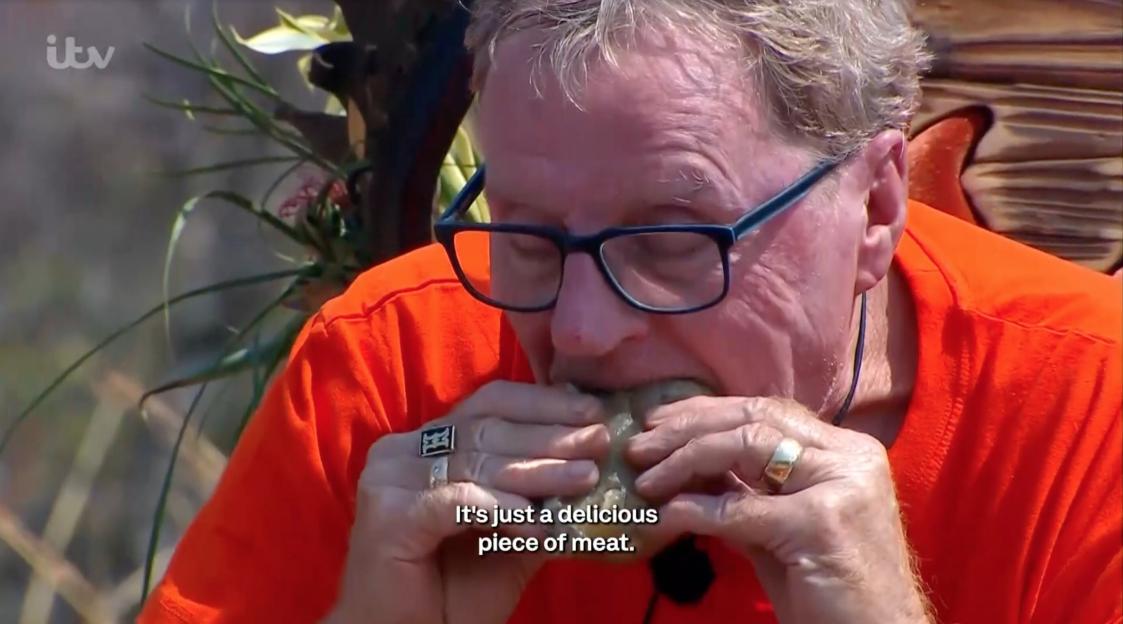 A man with glasses and a bright orange shirt eats a grayish piece of food.